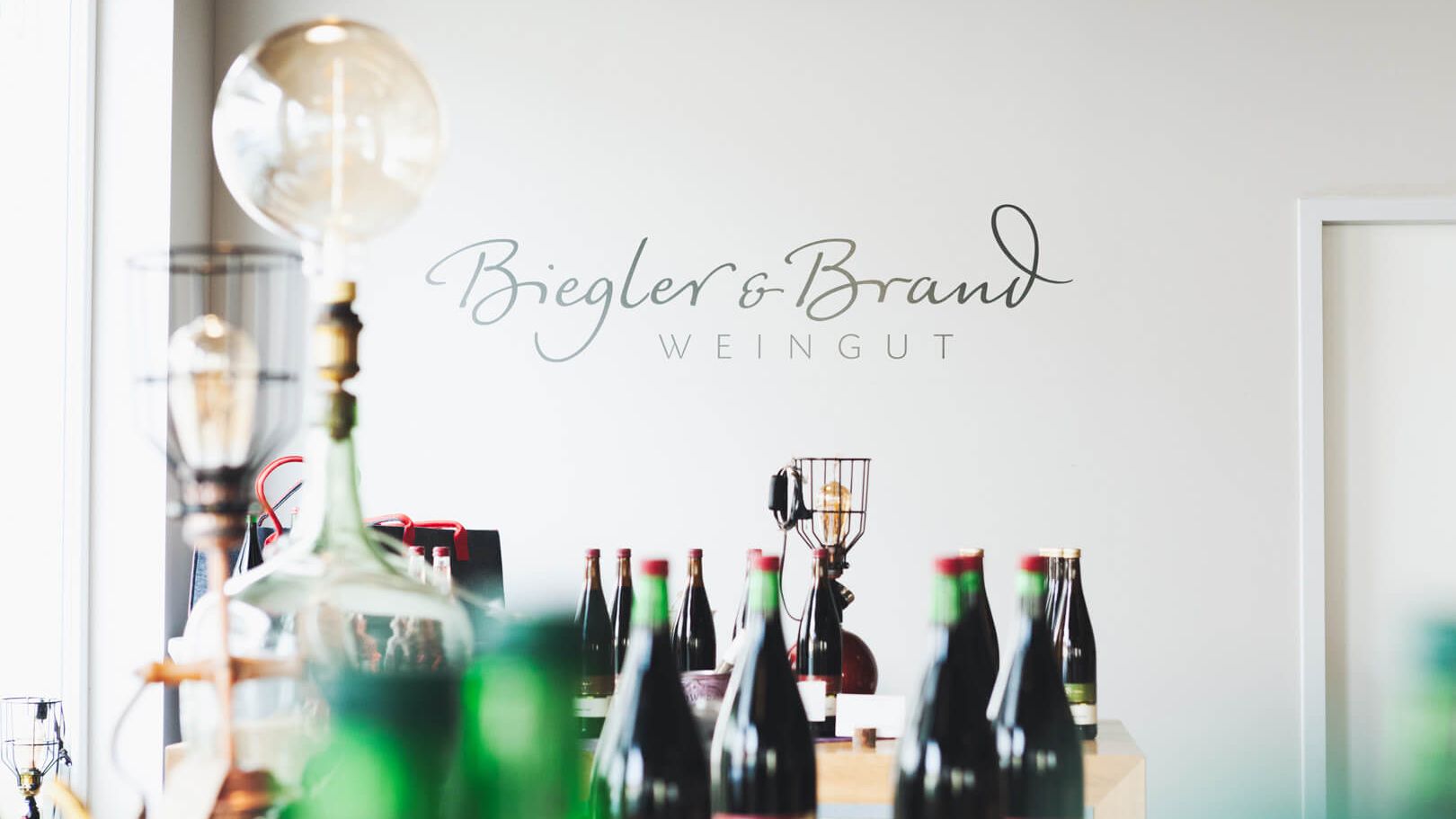 Verschiedene Weine in Flaschen im Weinladen des Weinhotel Kaisergarten Alzey mit Logo des Weinguts Biegler & Brand im Hintergrund